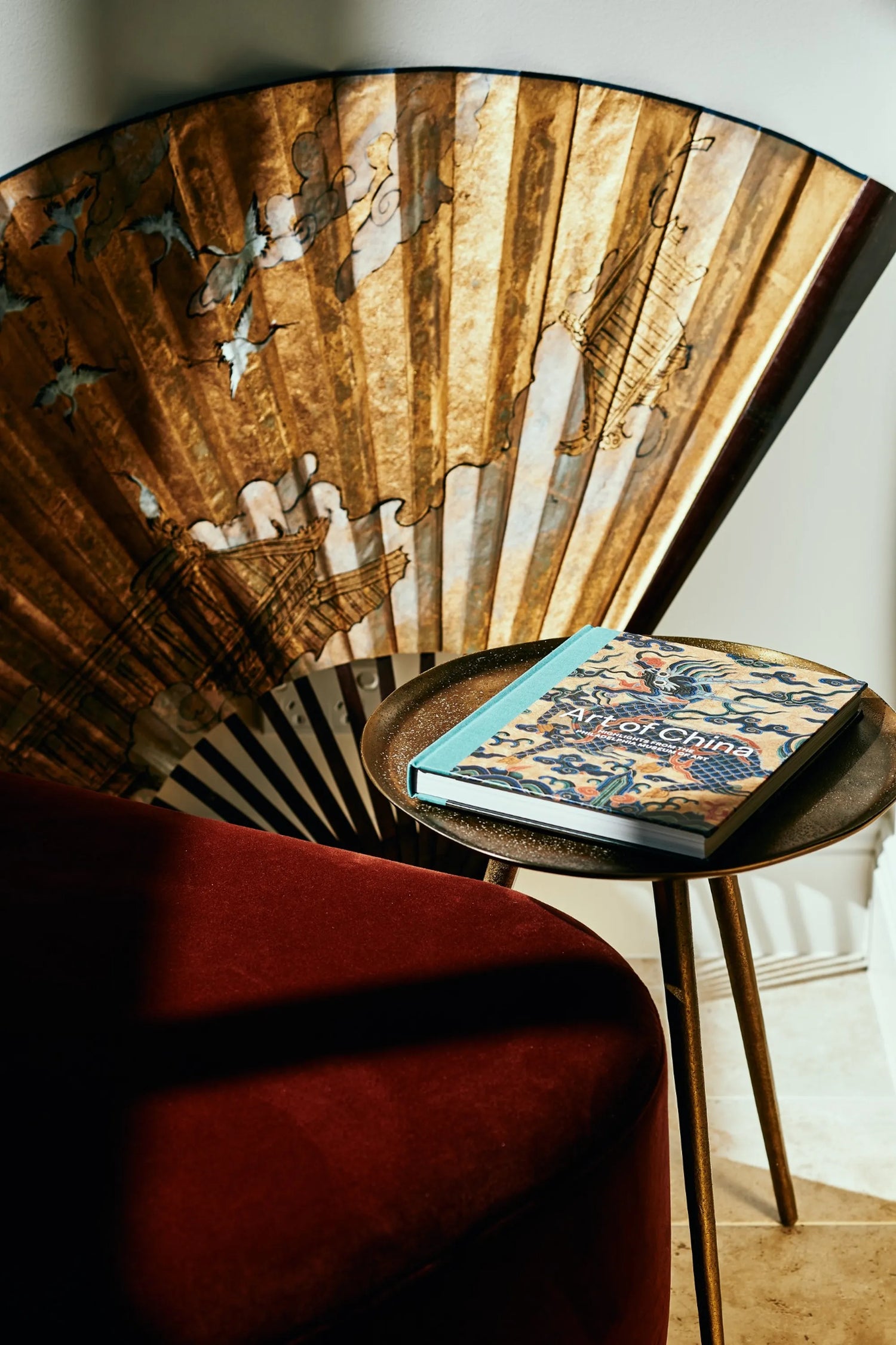 Decorative interior detail with Art of China book on round side table, antique golden fan backdrop and red velvet sofa