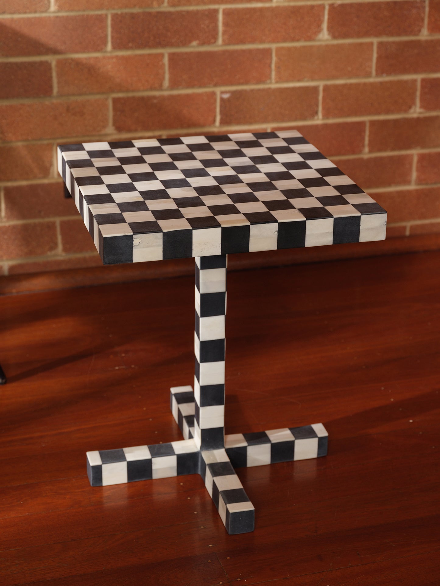 Modern black and white checkerboard side table with square top and pedestal base with brick wall background