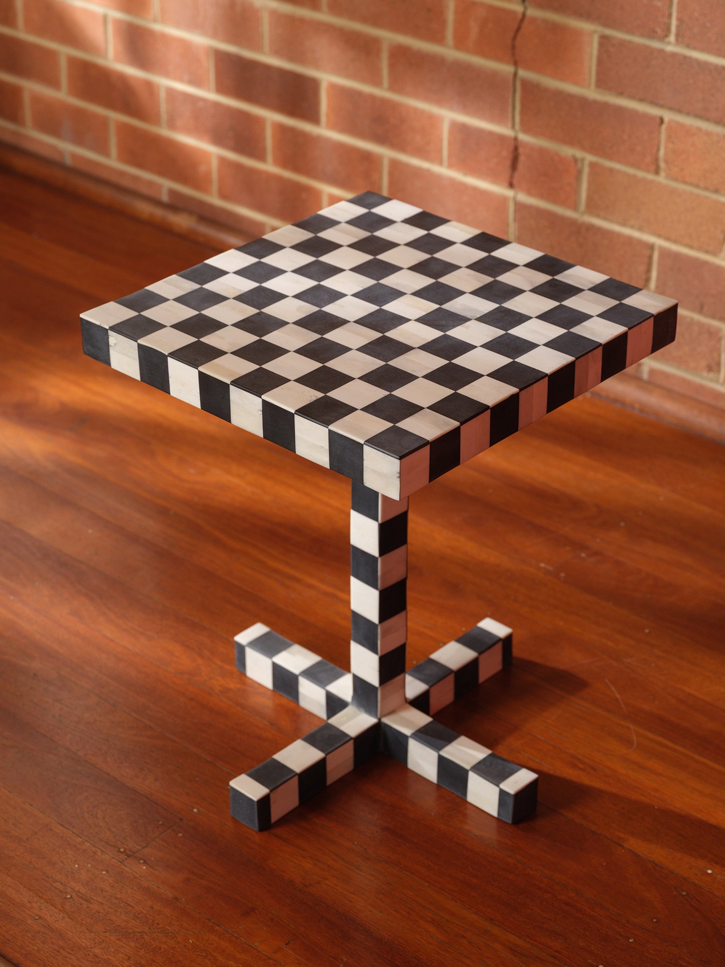 Modern black and white checkerboard side table with square top and pedestal base with brick wall background