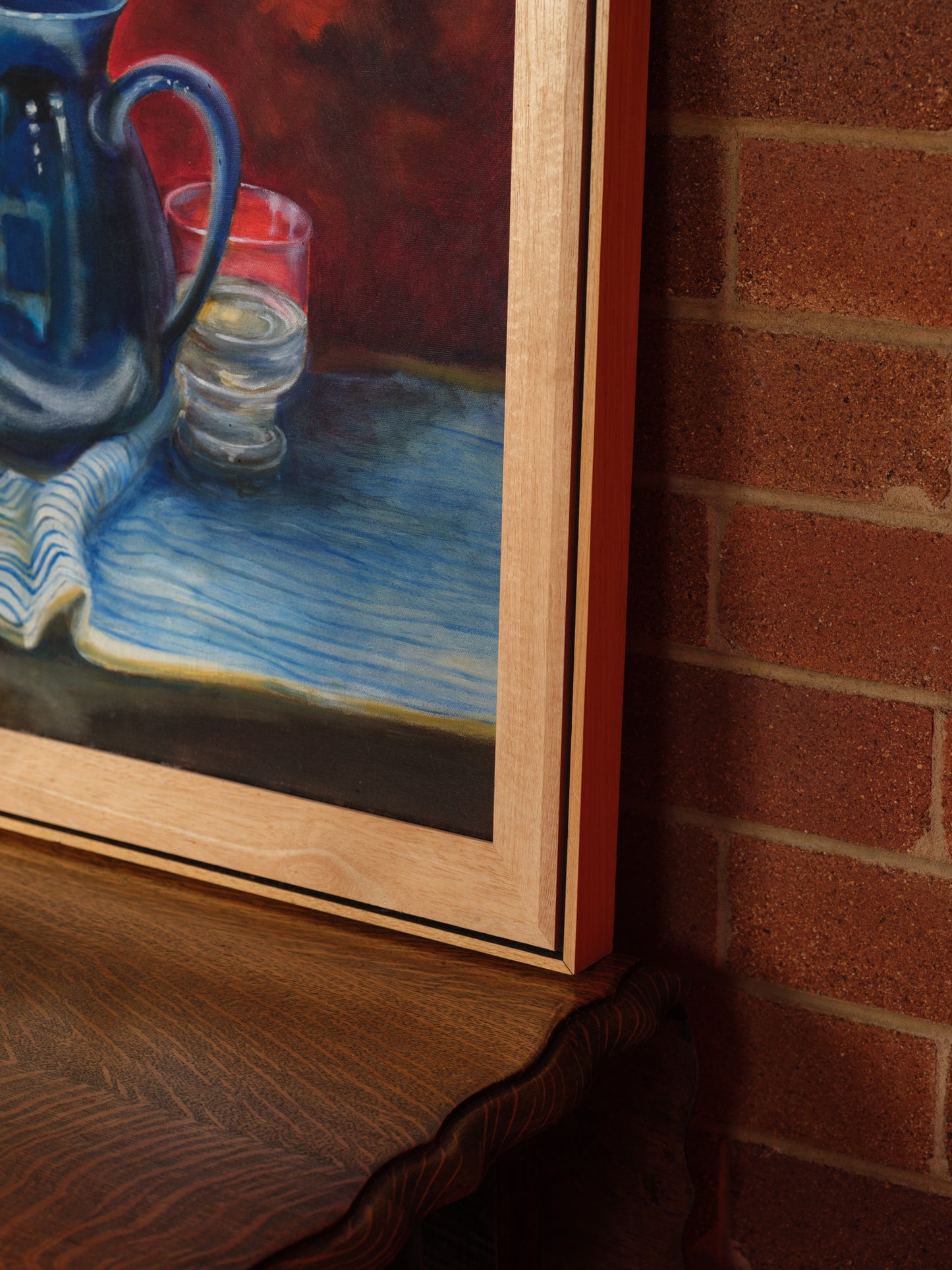 Oil painting titled Cobalt Pitcher and Glasses, featuring a deep blue jug and six clear glasses arranged on a striped cloth with a rich red background.