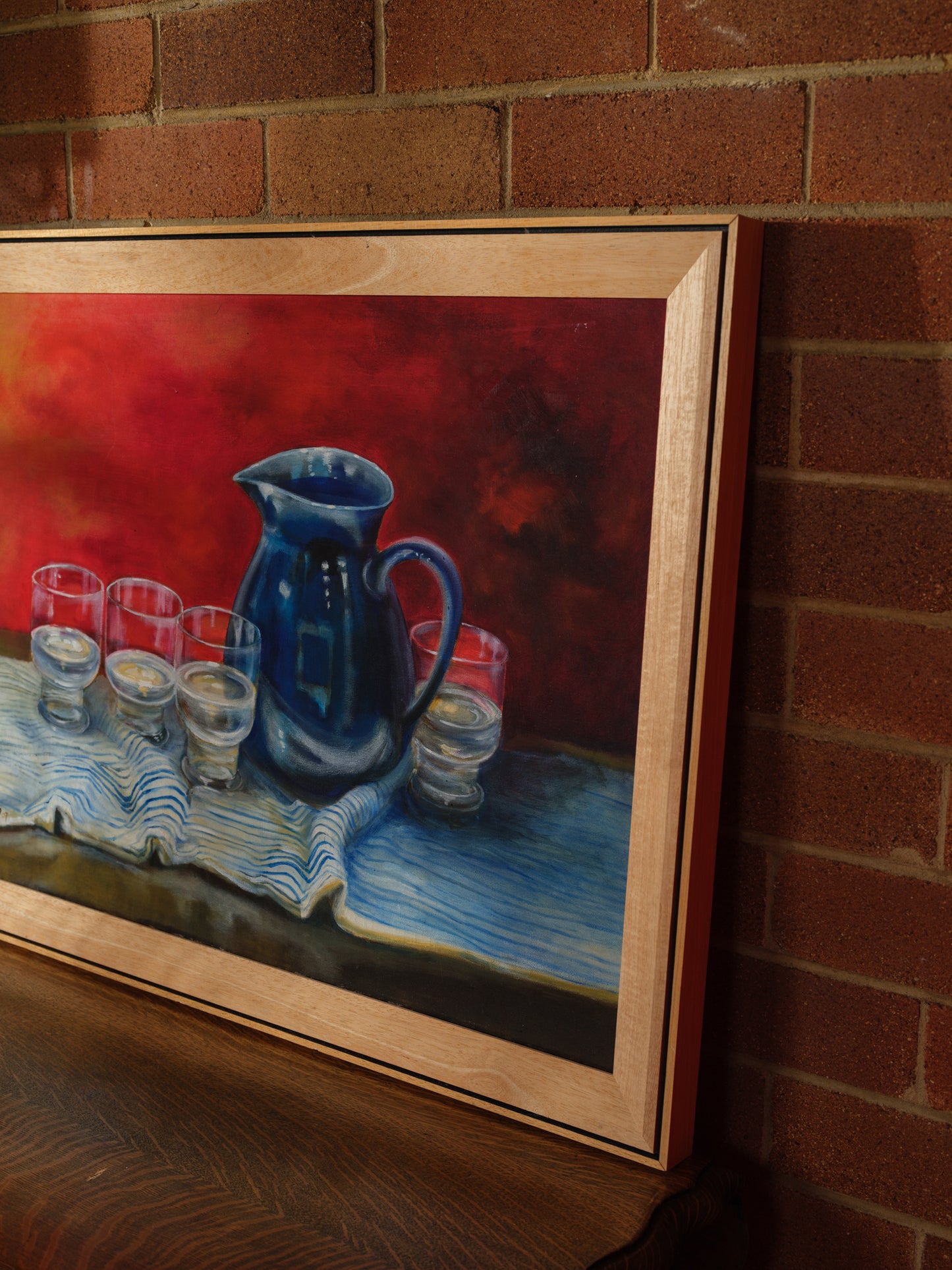 Oil painting titled Cobalt Pitcher and Glasses, featuring a deep blue jug and six clear glasses arranged on a striped cloth with a rich red background.