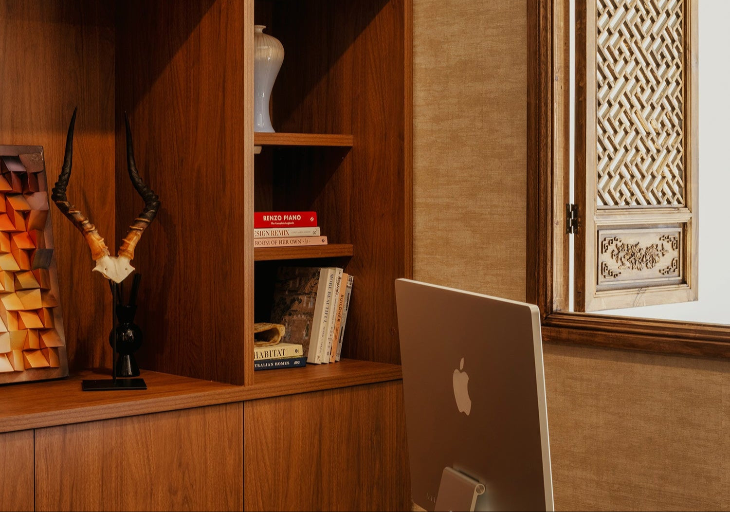 modern home office with wooden bookshelf, storage boxes, decorative items and iMac computer on glass desk beside traditional carved window