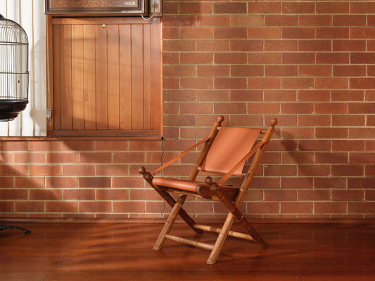 Replica faux bamboo and cognac leather safari style folding chair, vintage-inspired design, with brick wall