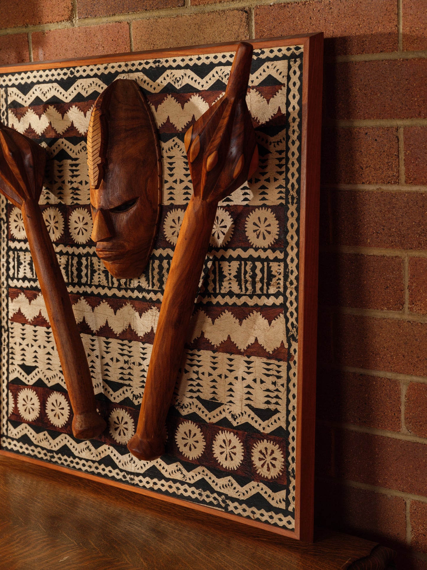 Carved hardwood mask relief artwork titled Tapa Guardian Mask Relief, featuring a central tribal mask and two stylized clubs mounted on traditional tapa bark cloth, set in a wooden frame.