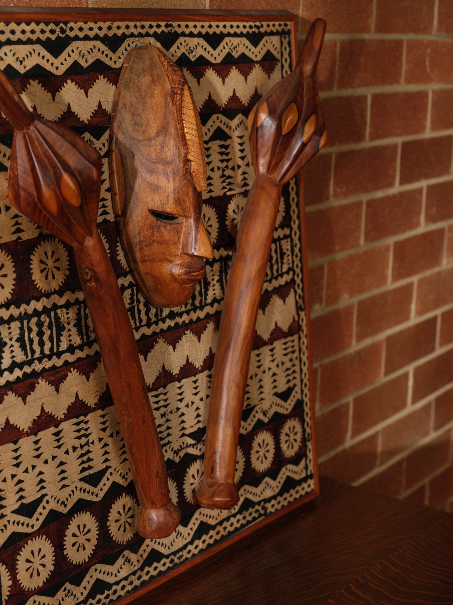 Carved hardwood mask relief artwork titled Tapa Guardian Mask Relief, featuring a central tribal mask and two stylized clubs mounted on traditional tapa bark cloth, set in a wooden frame.