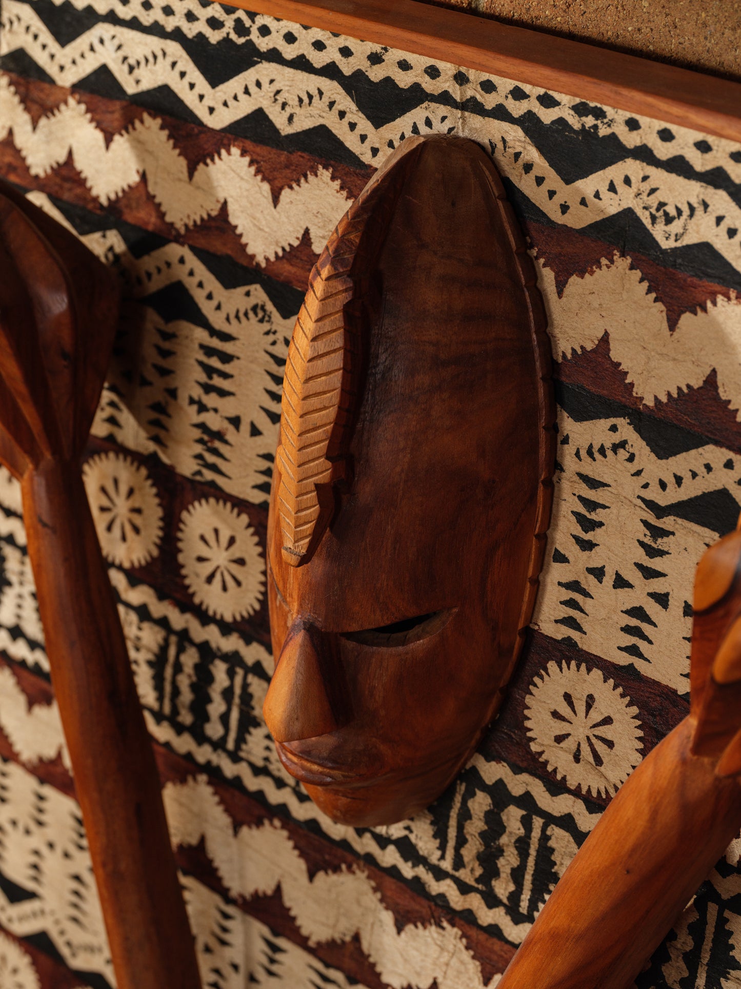 Carved hardwood mask relief artwork titled Tapa Guardian Mask Relief, featuring a central tribal mask and two stylized clubs mounted on traditional tapa bark cloth, set in a wooden frame.
