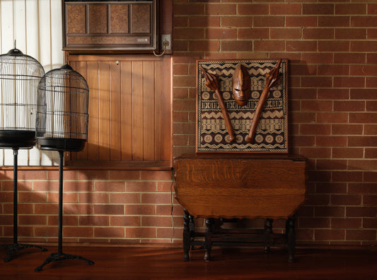 Carved hardwood mask relief artwork titled Tapa Guardian Mask Relief, featuring a central tribal mask and two stylized clubs mounted on traditional tapa bark cloth, set in a wooden frame.