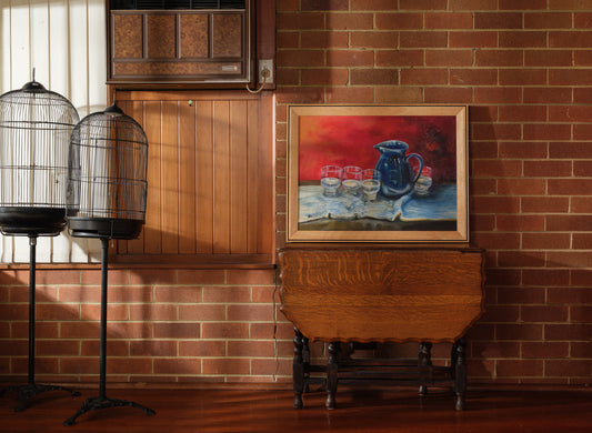 Oil painting titled Cobalt Pitcher and Glasses, featuring a deep blue jug and six clear glasses arranged on a striped cloth with a rich red background.