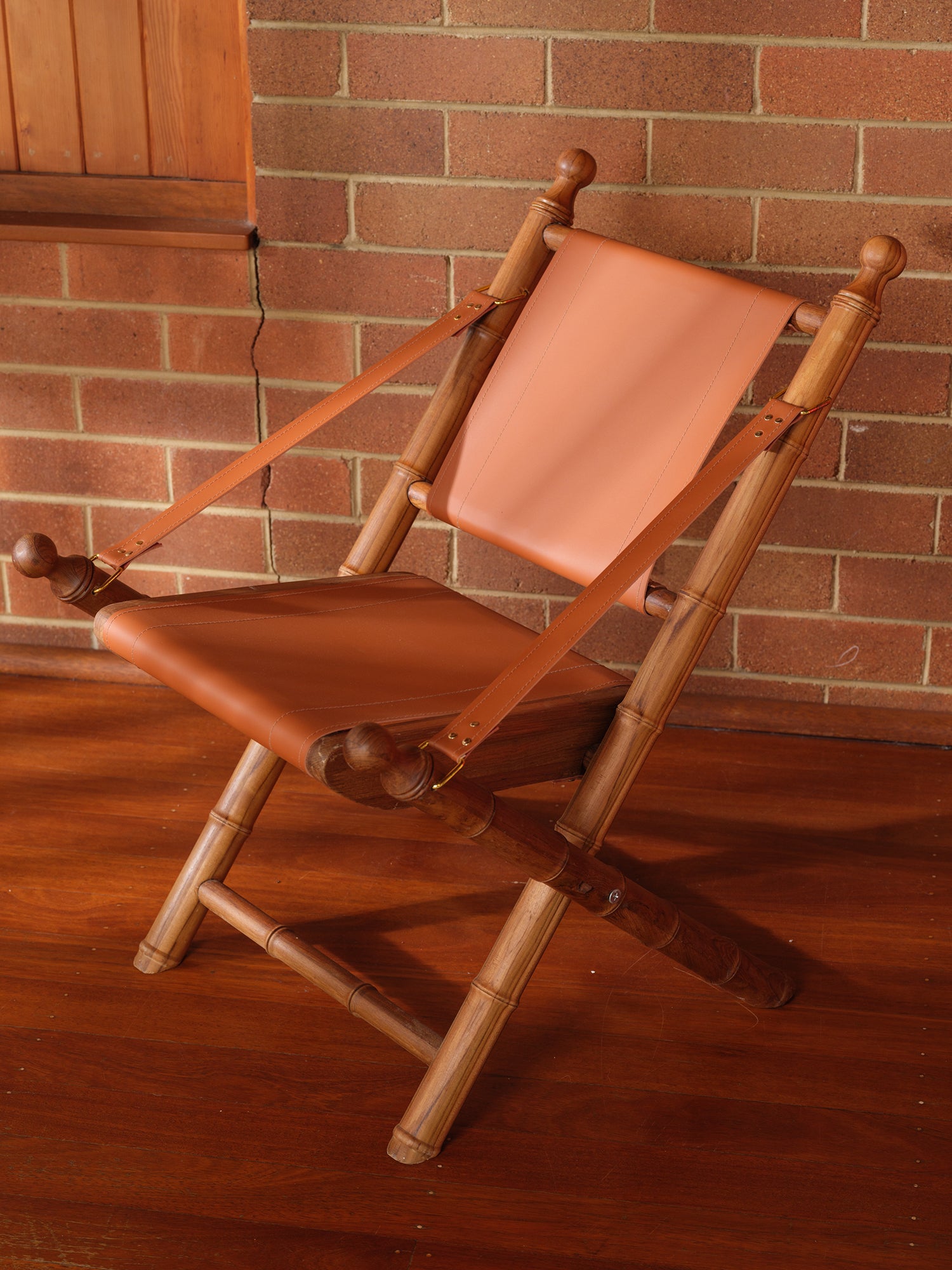 Replica faux bamboo and cognac leather safari style folding chair, vintage-inspired design, with brick wall
