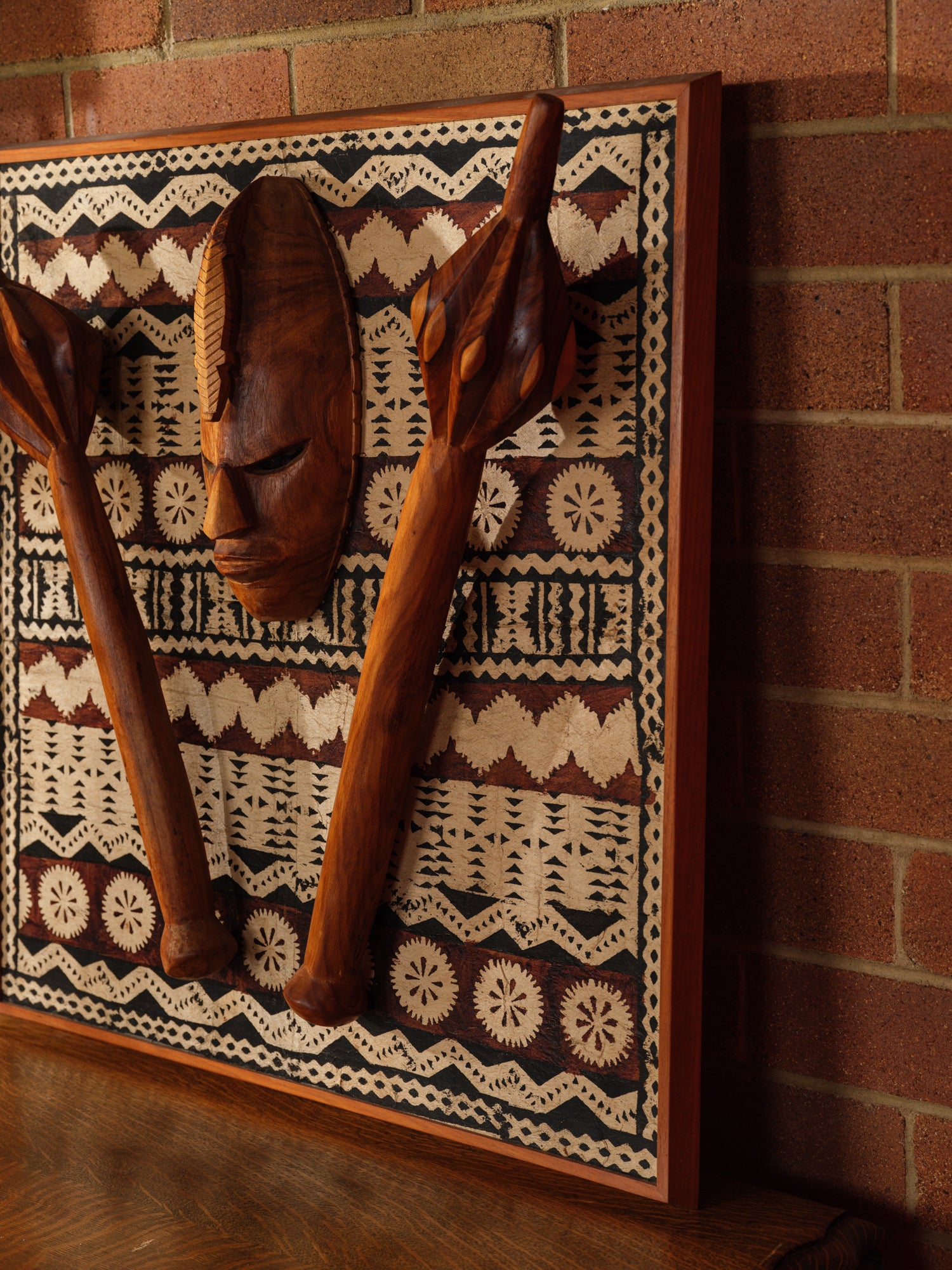 Carved hardwood mask relief artwork titled Tapa Guardian Mask Relief, featuring a central tribal mask and two stylized clubs mounted on traditional tapa bark cloth, set in a wooden frame.
