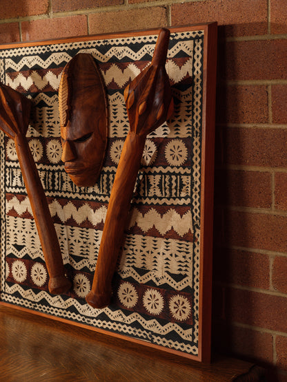 Carved hardwood mask relief artwork titled Tapa Guardian Mask Relief, featuring a central tribal mask and two stylized clubs mounted on traditional tapa bark cloth, set in a wooden frame.