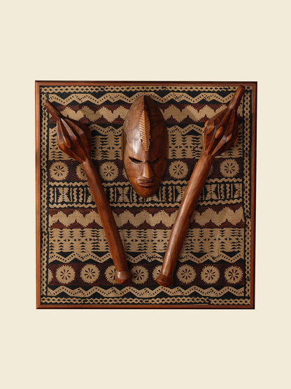 Carved hardwood mask relief artwork titled Tapa Guardian Mask Relief, featuring a central tribal mask and two stylized clubs mounted on traditional tapa bark cloth, set in a wooden frame.