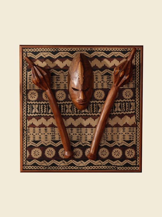 Carved hardwood mask relief artwork titled Tapa Guardian Mask Relief, featuring a central tribal mask and two stylized clubs mounted on traditional tapa bark cloth, set in a wooden frame.
