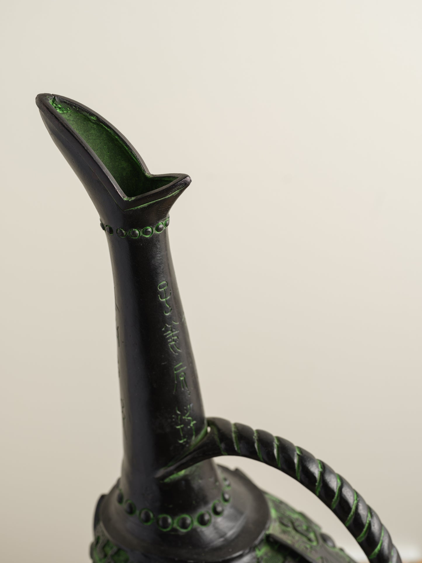Bronze-inspired black and green tripod vessel with tall spout and arched handle.
