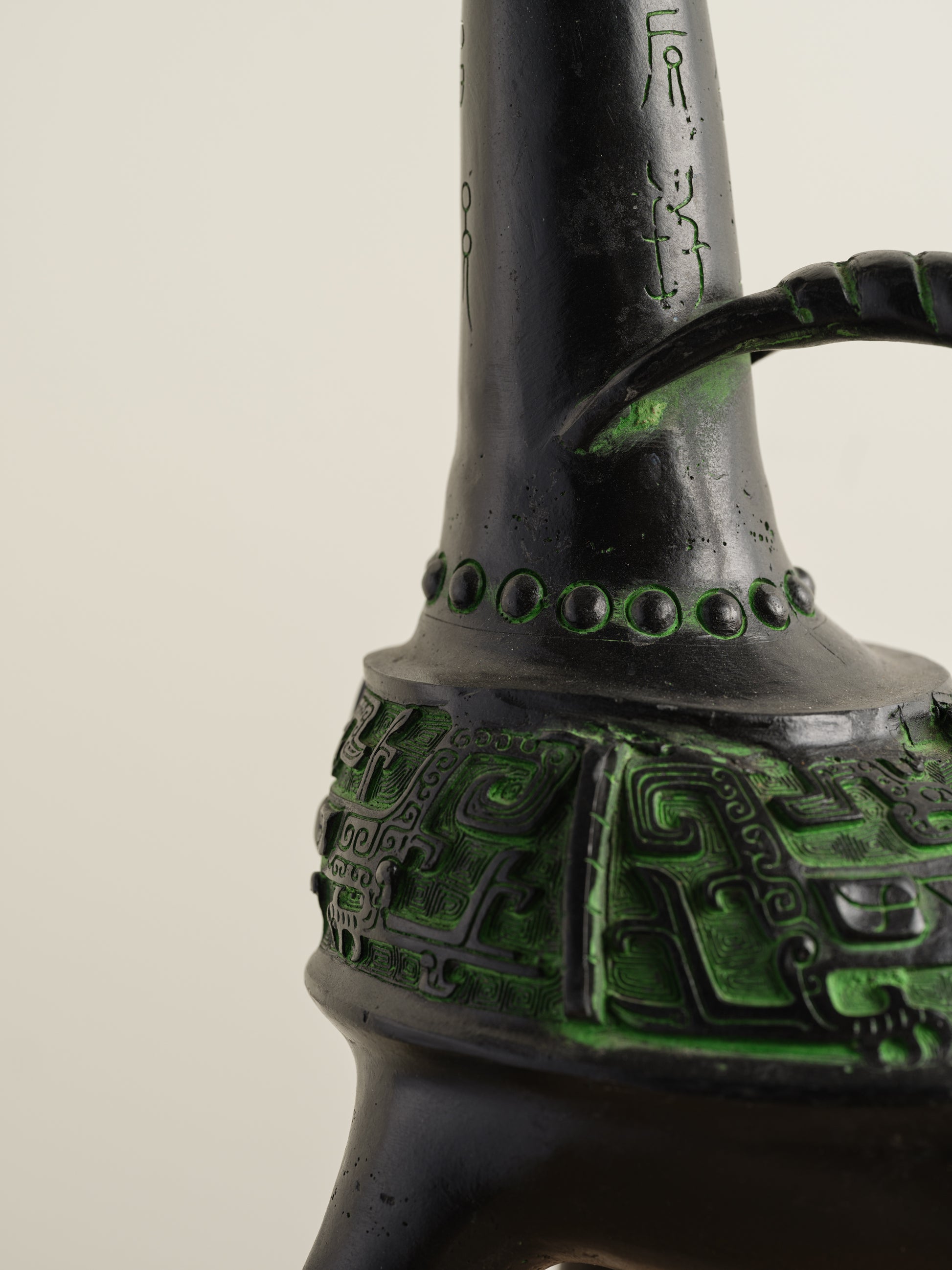 Bronze-inspired black and green tripod vessel with tall spout and arched handle.
