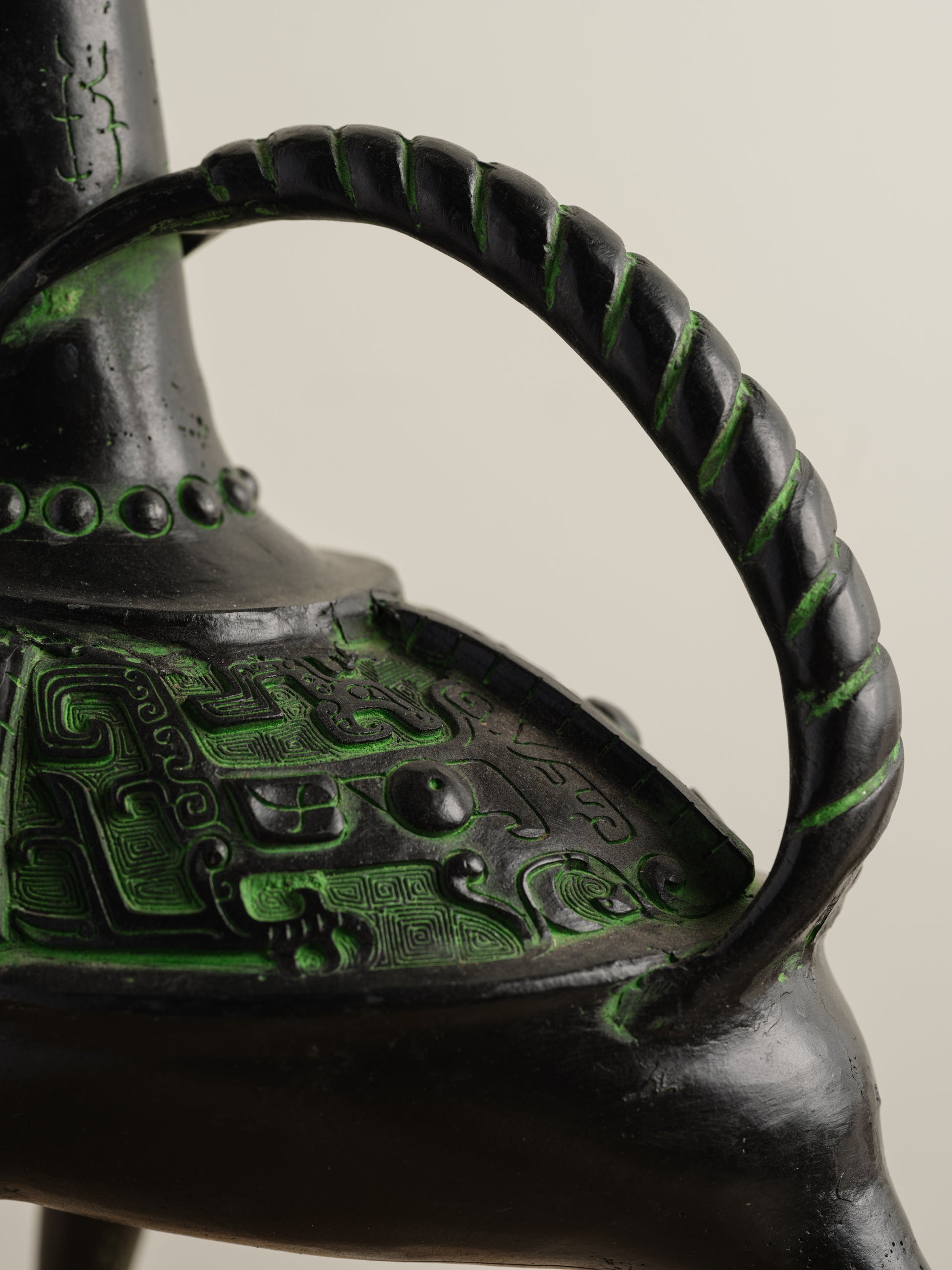 Bronze-inspired black and green tripod vessel with tall spout and arched handle.
