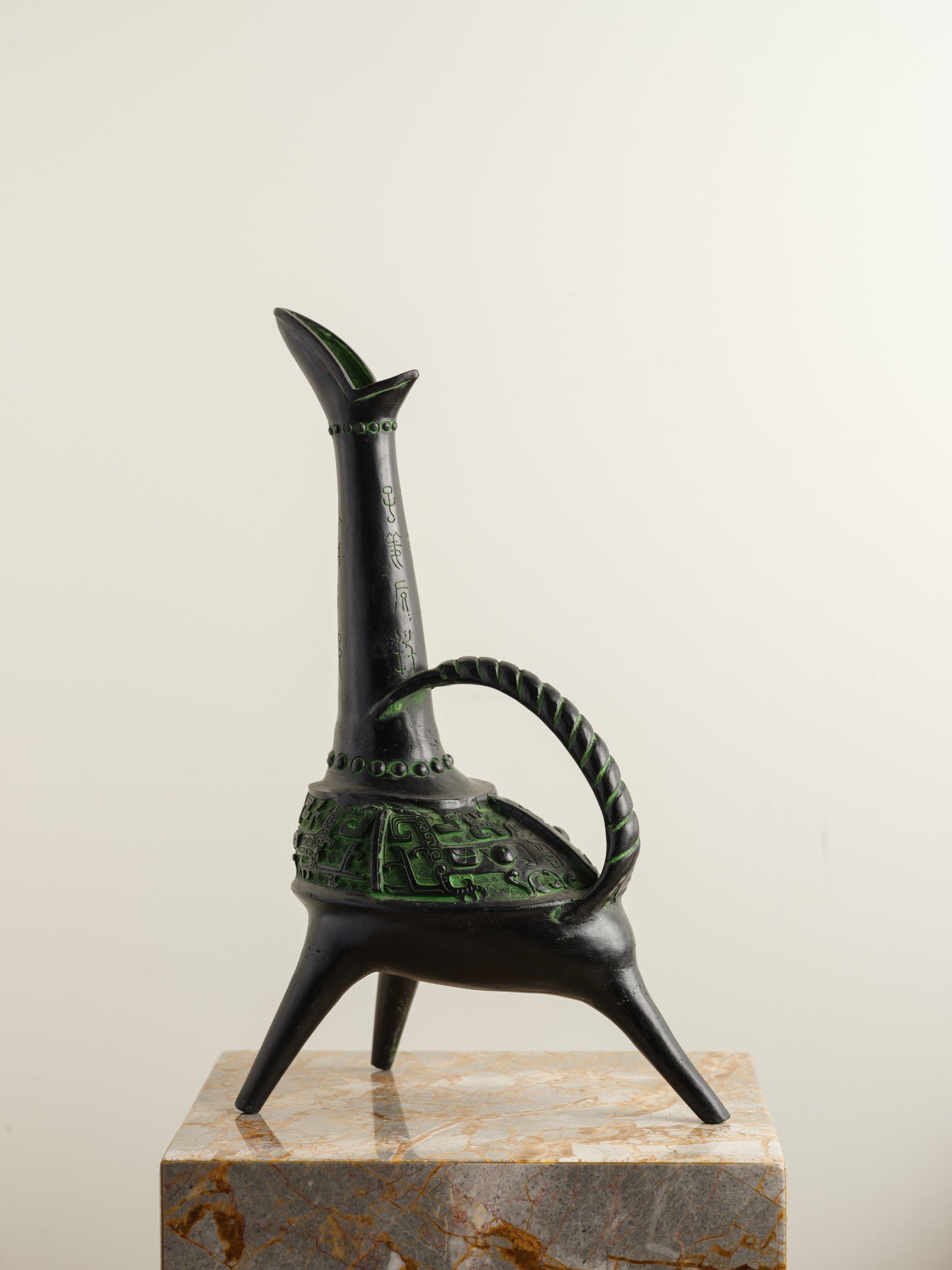 Bronze-inspired black and green tripod vessel with tall spout and arched handle.
