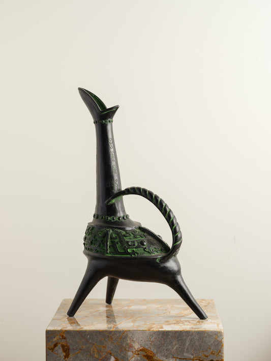Bronze-inspired black and green tripod vessel with tall spout and arched handle.
