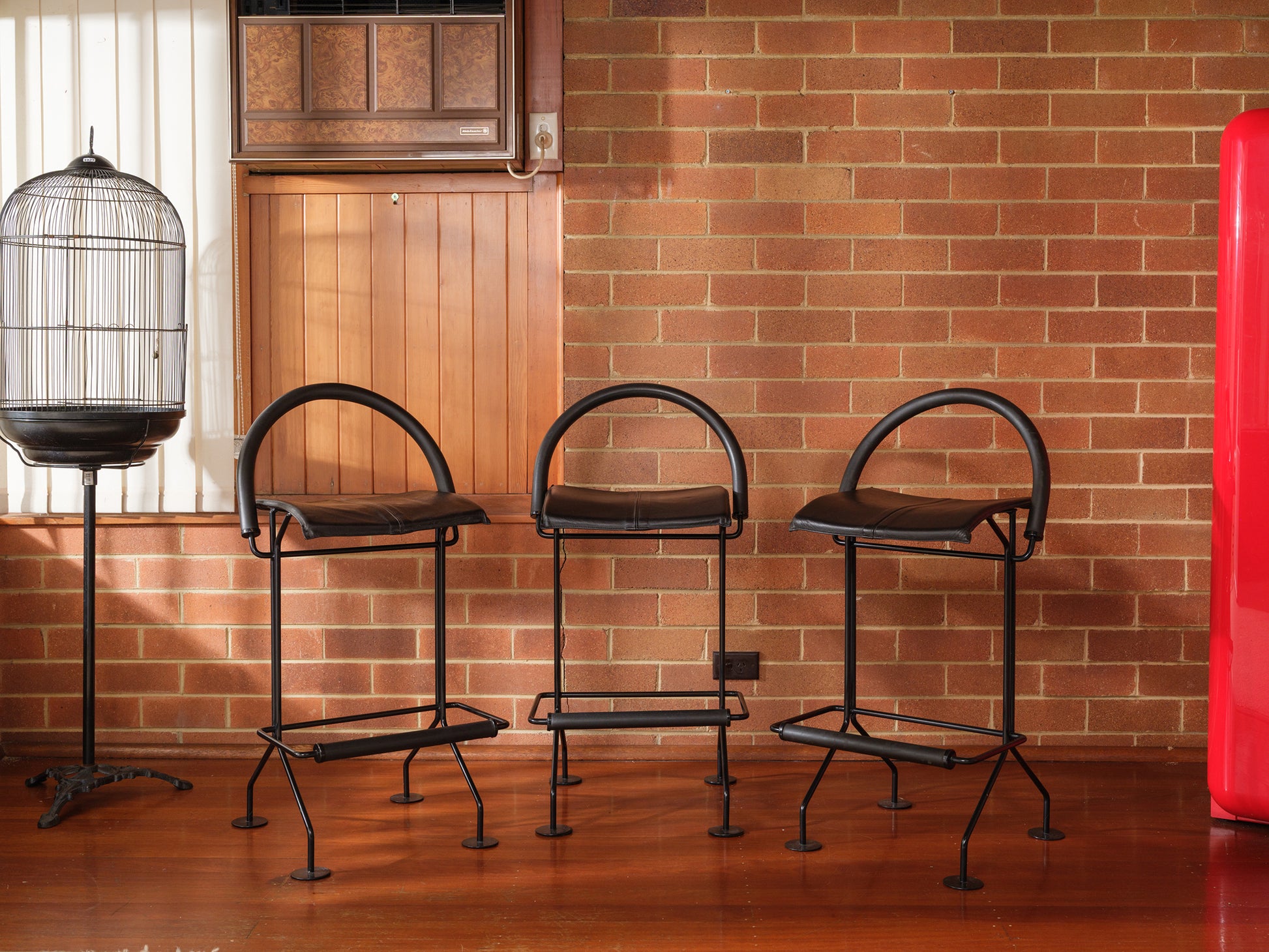 Three black bar stools against a brick wall with a birdcage and red object in the background.